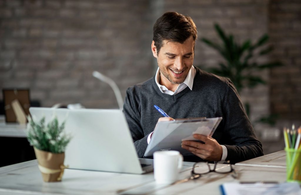 Empresário sorridente escrevendo relatório de negócios enquanto trabalhava no laptop no escritório