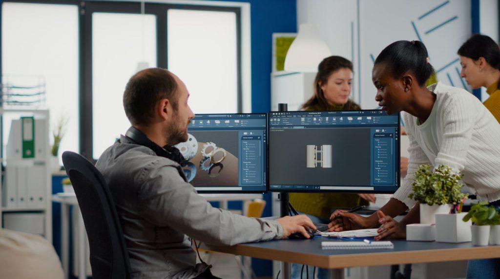 Dois funcionários, uma mulher negra e um homem branco, analisando peças metálicas mecânicas em 3D em monitores em um ambiente empresarial.