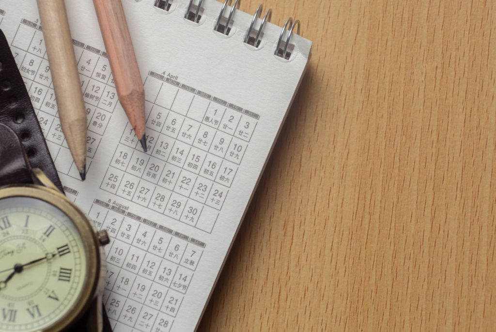 Agenda de papel, dois lápis e um relógio de pulso sobre mesa de madeira