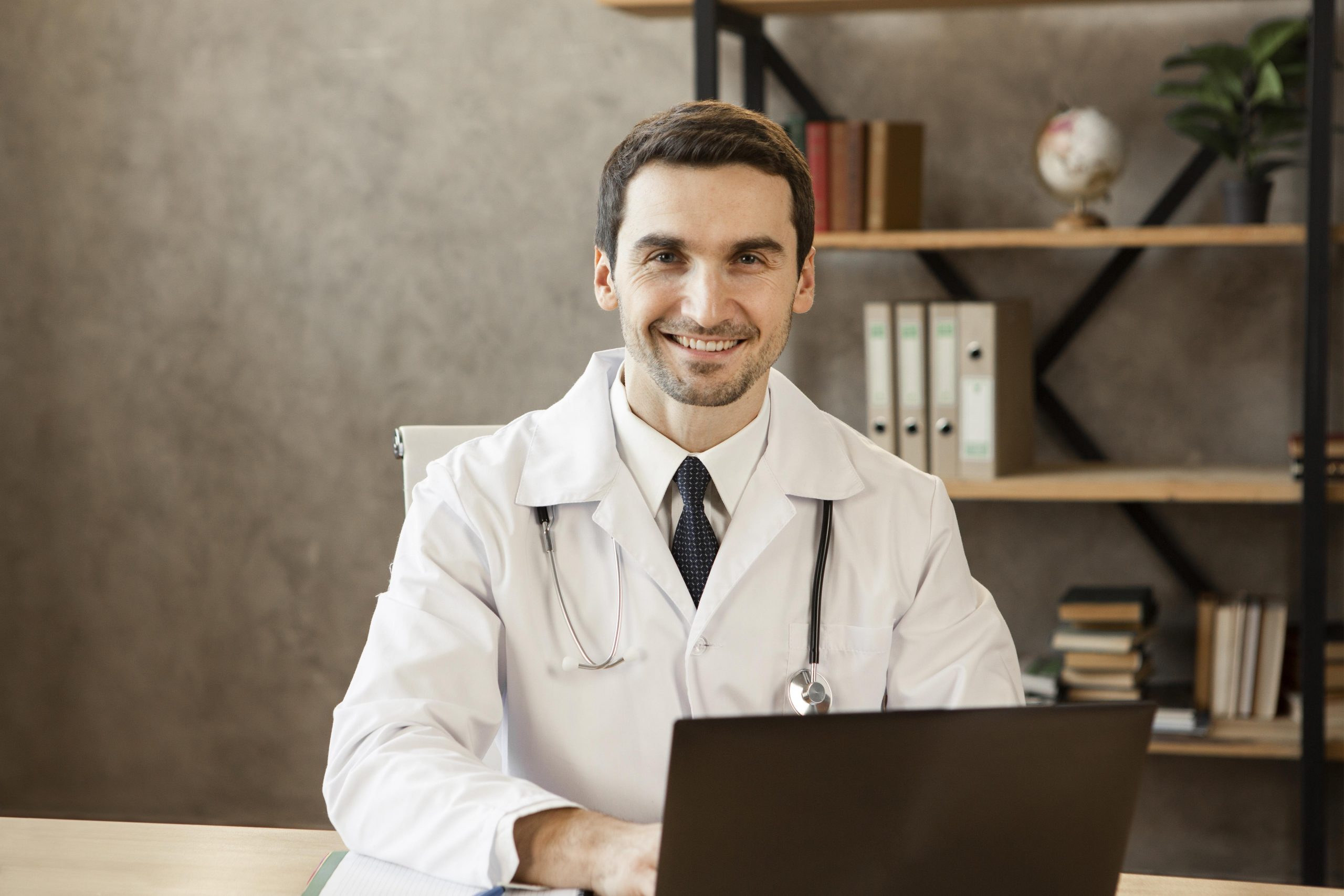 Médico jovem com jaleco e estetoscópio pendurado no pescoço, sentado em escritório e usando um notebook