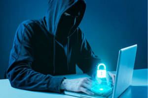 Homem encapuzado representando um hacker utiliza um notebook, no qual aparece um símbolo virtual de cadeado representando a segurança da informação