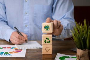 Homem em trajes formais empilha três blocos de madeira sobre a mesa, cada um com um símbolo que representa a sustentabilidade