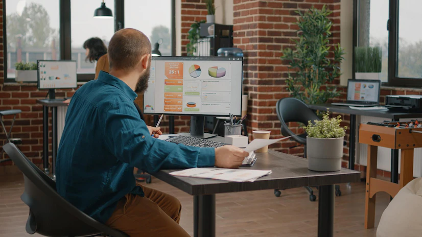 Homem sentado em seu escritório de frente para a tela de um computador segurando documentos