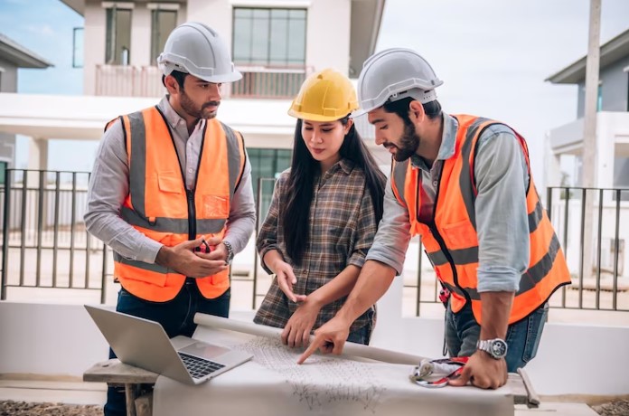 Três profissionais da construção civil usando capacetes e coletes de segurança trabalhando juntos no canteiro de obras construindo casas no conceito de trabalho em equipe e cooperação.