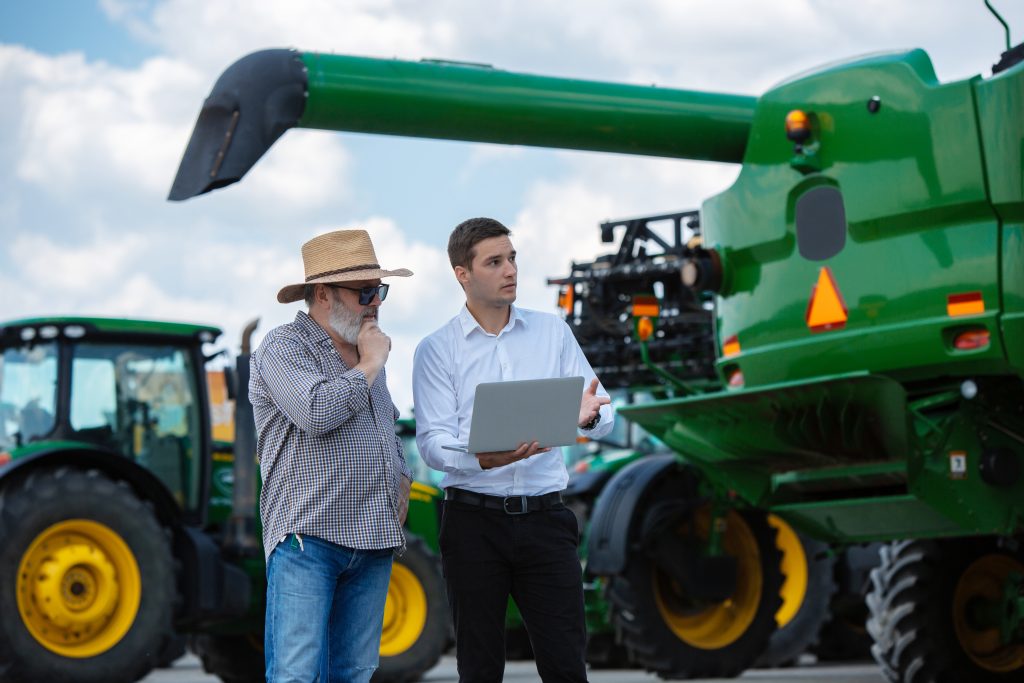 Dois empresários do agronegócio consultando dados em um notebook. Máquinas agrícolas ao fundo