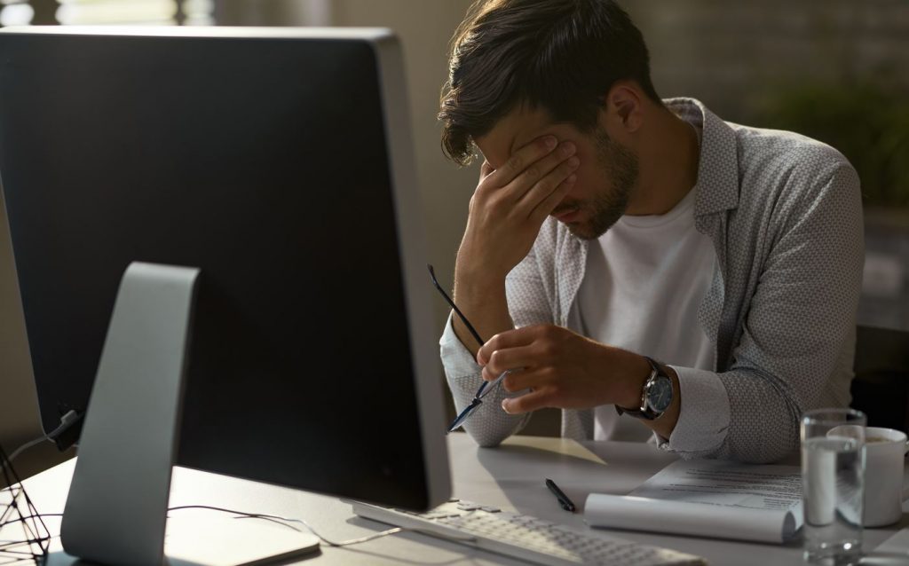 Homem exausto segurando a cabeça com dor enquanto trabalhava com gerenciamento de contratos no pc desktop em escritório