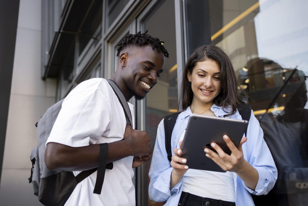 Dois jovens estudantes em ambiente acadêmico sorrindo enquanto olham para a tela de um tablet