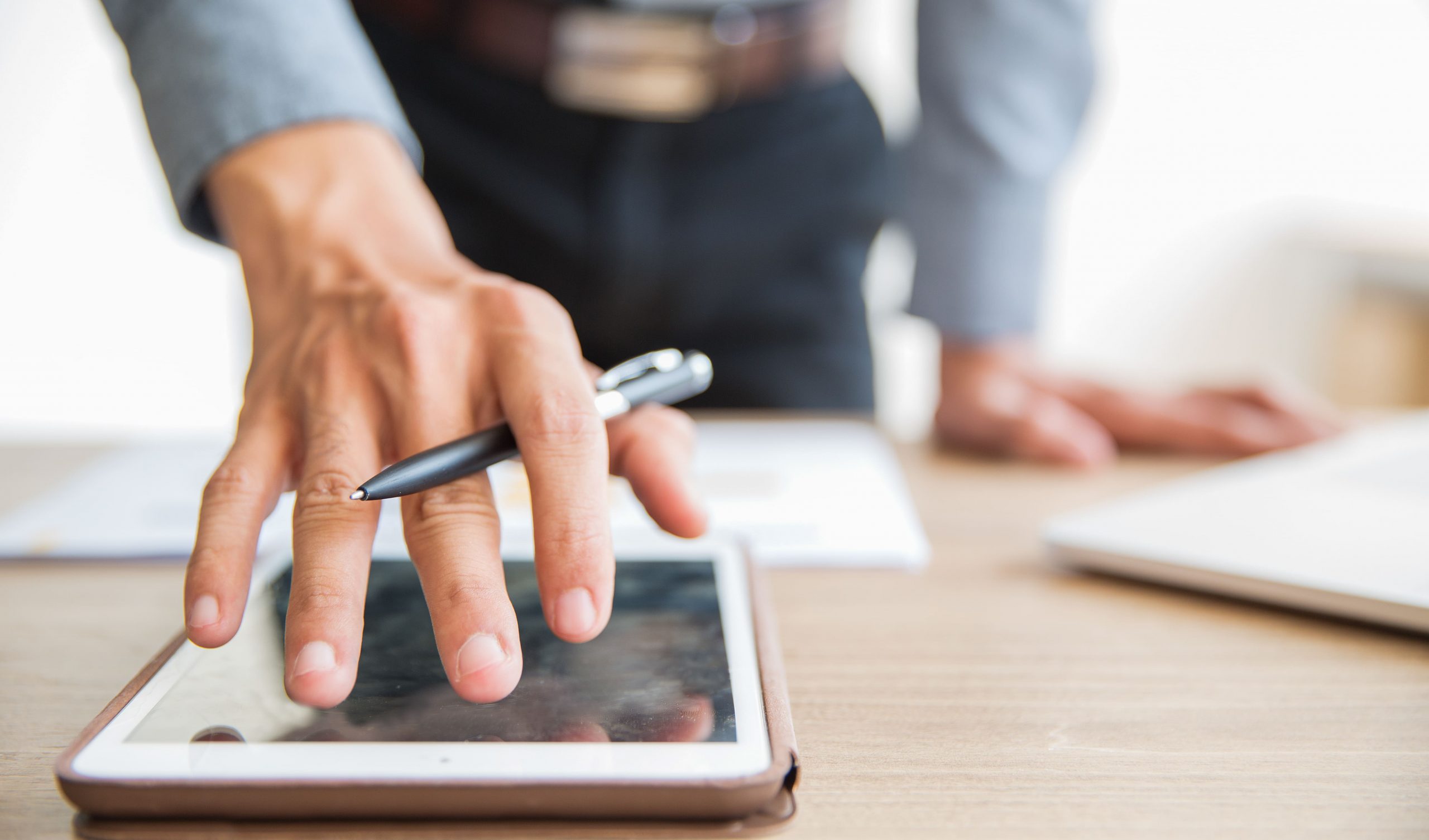 Mão de um homem executivo tocando a tela de um tablet enquanto segura uma caneta, representando uma assinatura digital. O dispositivo eletrônico está sobre uma mesa de madeira junto a outros objetos, como uma folha de papel e um notebook no canto direito.