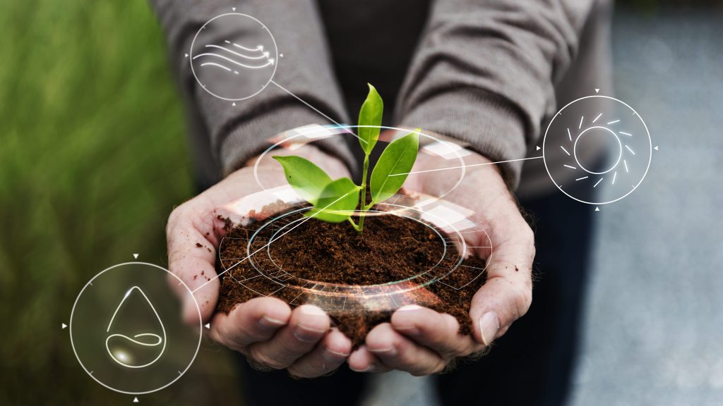 Imagem ilustra agricultura inteligente e rastreabilidade no agro. Duas mãos seguram e apresentam um punhado de terra com planta brotando. A frente, gráficos ilustram água, sol e ár, componentes do plantio.